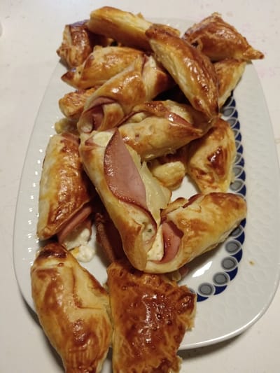 A close-up, overhead view depicts a white oval ceramic platter filled with approximately twenty irregularly shaped, golden-brown baked pastries. Many pastries exhibit a glossy finish, suggesting an egg wash. Several items reveal fillings: slices of pink, cooked ham are visible, often folded around a white, creamy substance that appears to be cheese, possibly with flecks of green herbs. Some pastries are crescent-shaped, while others are more triangular or rolled. The platter features a decorative border on its right side, consisting of interlocking dark blue and light gray semicircular or scalloped patterns. The platter rests on a light-colored, possibly white or off-white, textured surface. The scene is situated in Offenbach an der Queich, Germany.