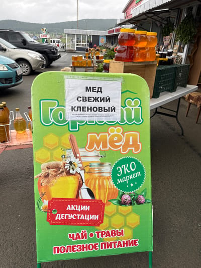 This is an eye-level, medium shot of a bright green, double-sided A-frame sign advertising honey products in Inzer, Russia. The sign is wet, suggesting recent rain. The foreground is dominated by the sign, which features large, colorful text and images. The text on the sign includes "МЕД СВЕЖИЙ КЛЕНОВЫЙ" (Honey Fresh Maple), "Горный мёд" (Mountain Honey), "ЭКО маркет" (Eco Market), "АКЦИИ ДЕГУСТАЦИИ" (Promotions Tastings), "ЧАЙ ТРАВЫ" (Tea Herbs), and "ПОЛЕЗНОЕ ПИТАНИЕ" (Healthy Nutrition). Images on the sign depict jars of honey, a bee, a spoon, and honeycomb patterns.

In the background, several cars are parked on a wet asphalt surface. A black SUV is prominent on the left. Behind the cars, a building with a red roof and white walls is visible, possibly a cafe or market stall. Further in the distance, mountains are faintly visible under an overcast sky. The overall setting appears to be an outdoor market or roadside stall.