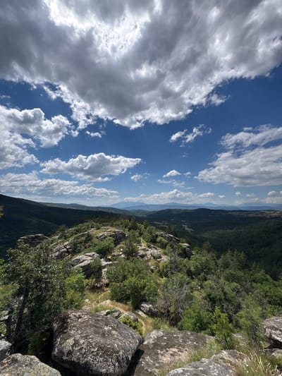 The image captures a scenic overlook of a mountainous landscape, likely situated in or near Dolno Drianovo, Bulgaria. The foreground is dominated by large, weathered boulders covered in lichen, interspersed with sparse green vegetation and dry grasses. Beyond these rocks, the terrain slopes downwards, covered in a dense forest of dark green trees. Further in the distance, rolling hills fade into the horizon, with a hazy, lighter green and blue hue suggesting a considerable distance and atmospheric perspective. The sky above is a vibrant blue, punctuated by dramatic, white cumulus clouds that cast shadows on the landscape below. The lighting suggests a bright, sunny day, possibly mid-afternoon, given the contrast between the illuminated clouds and the shaded parts of the terrain. No people or man-made structures are visible in the frame.