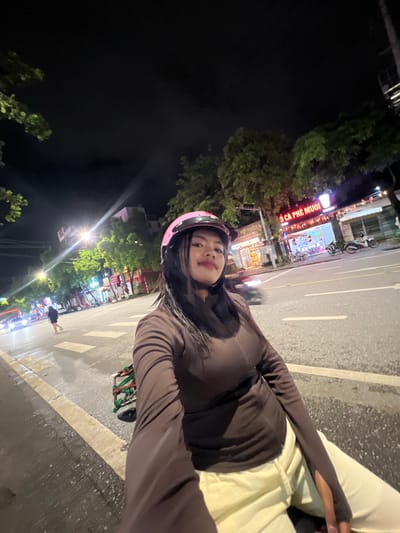 A young woman wearing a pink helmet and a brown jacket is taking a selfie on a motorcycle at night. The street is wet, suggesting it might have recently rained. In the background, there are shops and trees, with streetlights illuminating the scene. One shop sign in Vietnamese reads "BÒ CA PHE MUOI," indicating it is a coffee shop. Cars and motorcycles can be seen on the street. The woman is looking directly at the camera, with her hair wet.