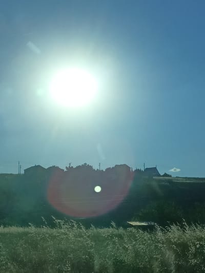 The image captures a rural scene bathed in bright sunlight. The sun is prominently positioned in the upper left quadrant, creating lens flares and a hazy atmosphere. In the middle ground, a silhouette of a rural landscape is visible. This includes several buildings, possibly houses or farm structures, scattered along a gentle slope. Trees and vegetation dot the hillside, and power lines can be seen stretching between poles. In the foreground, tall, wispy grass or reeds are illuminated by the sun. The overall impression is of a bright, sunny day in a countryside location, possibly Turkey given the location context of Bahşılı Belediyesi. There is no visible text in the image.