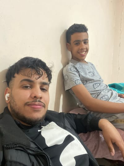 Two young men are lying on a light-colored floor. One is wearing a patterned gray shirt and smiling at the camera, his head resting directly on the floor. The other man is closer to the camera, wearing a black and white jacket, also lying on the floor with an earbud in his ear. The location is Sidi Chahmi, Algeria.