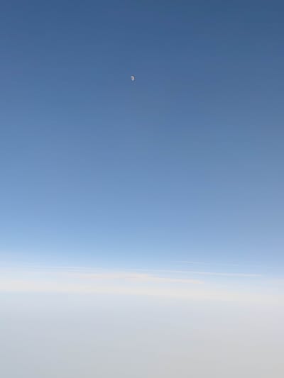 The image captures a vast expanse of clear, pale blue sky, transitioning to a lighter, almost white haze towards the bottom. A thin, wispy cloud formation stretches horizontally across the middle ground, appearing as a faint white line against the lighter sky. In the upper portion of the frame, a small, crescent-shaped moon is visible as a bright white sliver against the deep blue. The overall impression is one of a serene, high-altitude view, possibly from an airplane, given the atmospheric perspective and the presence of the moon in daylight. The lighting suggests it could be late afternoon or early morning, when the sky is still bright but the sun is not directly overhead. No people, objects, text, or specific location cues are discernible, beyond the general implication of being high above the earth's surface, over Novogrebenshchikovo, Russia.