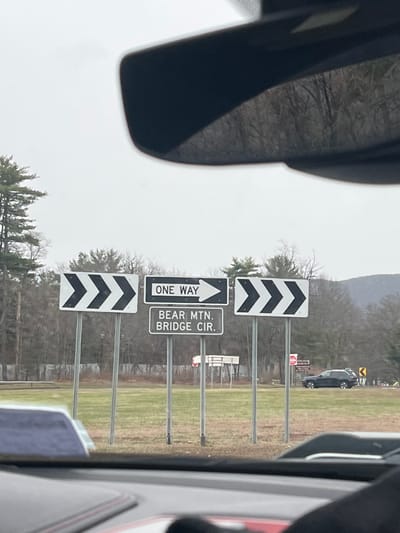 The scene depicts a daytime, overcast exterior view from inside a vehicle. In the midground, a cluster of four road signs stands on individual metal poles within a grassy median or roadside. From left to right, a white rectangular sign with black right-pointing chevrons is followed by a white rectangular sign with black text "ONE WAY" and a right-pointing arrow. Directly beneath this, another white rectangular sign displays "BEAR MTN. BRIDGE CIR." in black text. To the right of these, a third white rectangular sign with black right-pointing chevrons is visible. A dense line of leafless deciduous trees and some evergreen trees forms the background, with a distant mountain or large hill visible on the right under a light grey, overcast sky. A dark-colored SUV is present on a road to the right, behind the signs. The location is an approach to the Bear Mountain Bridge Circle, within the Town of Cortlandt, New York, United States of America.