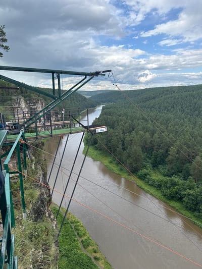 This media file captures a scenic view from a high vantage point overlooking a wide river and a dense forest. The scene appears to be a recreational area, possibly a bungee jumping or ziplining site, given the prominent metal structures and cables. The foreground features a robust green metal frame, suggesting a platform or support for an activity. Multiple cables, some orange and some black, stretch across the frame, disappearing into the distance over the river and forest.
The river flows through a valley, with steep, tree-covered slopes rising on the opposite bank. The forest is a lush, deep green, indicating it is likely summer or late spring. The sky above is dynamic, with dramatic, billowing clouds that are a mix of white and grey, suggesting an overcast or partly cloudy day. The lighting indicates it's daytime, but the heavy cloud cover might be tempering the sunlight.
In the middle ground, a platform with green artificial turf is visible, along with more metal structures and netting, further supporting the idea of an adventure activity. A white sign with some text is attached to a pole on the platform. There is no discernible activity of people, but the infrastructure suggests people are expected or have been using the area. The overall mood is one of natural grandeur combined with man-made adventure structures.
One visible sign on the platform has the text "Gravity" and "100 M", likely indicating a height measurement or the name of the attraction. The location is specified as Kul'metovo, Russia.