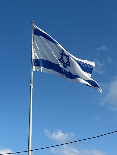 The image shows the flag of Israel flying against a bright blue sky with scattered white clouds. The flag is white with two horizontal blue stripes and a blue Star of David in the center. It is attached to a tall, silver flagpole. The flagpole appears to be located in an outdoor setting, possibly a public space. The image was taken in Kfar Shmaryahu, Israel. There are no people or animals visible in the image. There is a thin black wire running horizontally across the lower part of the frame, obscuring a small portion of the sky.