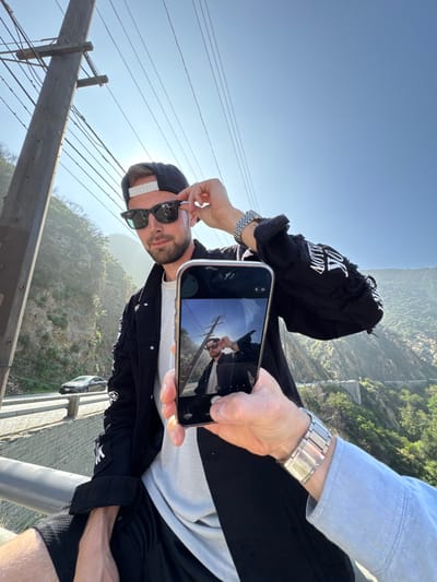 A forensic scene analysis depicts a sunny outdoor setting in the Unincorporated Santa Monica Mountains, United States of America. A male subject, appearing to be in his 20s or 30s, with short light brown hair and facial stubble, is positioned centrally. He wears a black backward baseball cap with a white strap, black Wayfarer-style sunglasses, a white t-shirt, a distressed black denim jacket with white text and graphic elements, and black shorts. A silver-toned metal link wristwatch is on his left wrist. He is seated or leaning against a concrete barrier, looking directly at the camera, with his right hand adjusting his sunglasses.
In the immediate foreground, the right arm and hand of a second individual are visible, wearing a light blue long-sleeved shirt and a similar silver-toned metal link wristwatch. This individual holds a silver-framed smartphone, likely an iPhone, vertically, with its screen displaying a live camera preview of the primary male subject.
The background consists of steep, verdant mountain slopes under a clear, bright blue sky. A winding paved road is visible on the left side, below the primary subject, where a dark-colored sedan is present. A tall wooden utility pole with multiple overhead wires stands prominently on the left. Strong sunlight from the upper left creates a discernible sunburst effect.