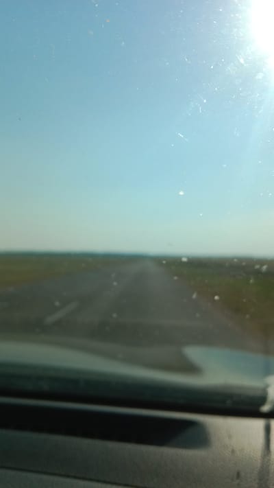 The image shows a view from inside a vehicle, looking out through a dusty windshield. Ahead, a straight, paved road recedes into the distance, bordered by flat, grassy terrain. The sky is a pale blue, with the sun shining brightly in the upper right corner, casting a glare and highlighting numerous specks of dust or dirt on the windshield. The foreground is dominated by the blurred dashboard and windshield frame of the vehicle. The scene suggests a drive through a rural or open area, possibly in or around Мірскі сельскі Савет, Belarus, given the flat landscape and the type of road.