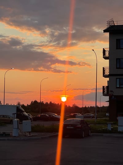 The image shows a sunset scene in the city of Poselok Kornevo, Russia. The sun is low on the horizon, casting a warm orange glow across the sky and illuminating the clouds with pink and orange hues. A prominent vertical beam of light, likely caused by atmospheric conditions or lens flare, cuts through the center of the frame.
In the foreground, there is a paved parking lot with several cars. One black sedan with a license plate reading "P723OC 178" is parked in the center. To the left, a white cargo van with its rear door open is visible. Adjacent to the van are utility boxes with electrical connections. Further to the left, a silver car is parked.
In the midground, a line of trees forms a dark silhouette against the vibrant sky. Several streetlights with curved poles are dotted throughout the scene, one of which is illuminated.
On the right side of the image, a multi-story brick building stands, featuring balconies with black railings and dark-tinted windows. A scaffolding structure is visible on the roof of the building. The overall environment suggests a residential or commercial area within Poselok Kornevo during the evening.