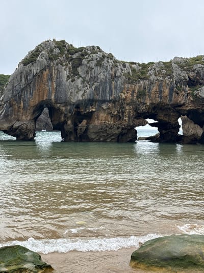 The image shows a natural rock arch formation in the sea at the beach of Poo, located in the municipality of Llanes, Spain. The arch is made of weathered gray and brown rock with patches of green vegetation on its surface. The sea is calm with clear, greenish water. Small waves are breaking on the sandy shore in the foreground, where a large, moss-covered rock is visible. The sky is overcast. There are no people or animals in the frame.