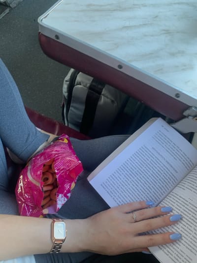 The image provides a close-up, high-angle view inside a public transport vehicle, likely a train or bus, located within the city of Vorokhta, Ukraine. The scene focuses on the lower body and left arm of an individual, dressed in grey leggings, seated beside a fixed table. This table features a light, marble-patterned surface, a red-brown padded edge secured by silver-colored rivets, and a light grey trim.

On the individual's lap, a bright pink plastic bag is open, revealing numerous small, ring-shaped dried bread products. The person's left hand, resting on their thigh, holds an open paperback book. The nails on this hand are manicured with light blue polish, and a silver-colored ring with a small clear stone is visible on the ring finger. A rose gold-toned rectangular watch with a metal link band is worn on the left wrist. The visible pages of the book display Cyrillic text, with the prominent heading "МЕНТАЛЬНОСТЬ ЗАВОЕВАТЕЛЕЙ" (MENTALITY OF CONQUERORS) clearly legible on the left page. In the background, partially visible behind the individual's legs, is a grey and black backpack or duffel bag, resting on a dark grey floor. The individual appears to be engaged in reading while consuming a snack.