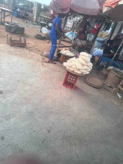 The image depicts a street scene in Gaga, Nigeria, likely a marketplace. A woman sits under a makeshift stall with a pink tarp, surrounded by goods. She appears to be selling "Kuli-Kuli," which are stacked high on platters atop a red plastic crate. A man in a blue tracksuit stands nearby, potentially a customer or vendor. Other individuals are seated in the background, suggesting a bustling market environment. Tires, plastic containers, and wooden structures indicate a semi-permanent or ad-hoc marketplace setup. The ground is a mix of dirt and concrete.