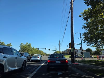 The image depicts a daytime scene on a sunny day, likely in Elk Grove, California. The perspective is from inside a vehicle, looking forward at a traffic intersection. Several cars are stopped in traffic, suggesting a queue or a red light.
In the foreground, the rear of a black sedan is visible, its taillights illuminated. The license plate reads "4AVH279". To the left, a white SUV is partially visible. Further ahead, other vehicles are stopped, including a silver sedan with license plate "5LNW808" and a dark-colored SUV.
The setting appears to be a suburban or commercial area with buildings and trees lining the road. A traffic light is visible, currently red for the lane the photographer is in. Utility poles with numerous wires stretch across the sky, and a street sign indicates "Laguna Blvd". On one of the utility poles, there is a poster for a "Reptile Expo". Another sign nearby advertises "DSV".
The overall mood is calm and ordinary, capturing a typical moment in urban traffic. The clear blue sky and bright sunlight suggest fair weather. The presence of traffic signals and multiple vehicles indicates an active road and intersection.