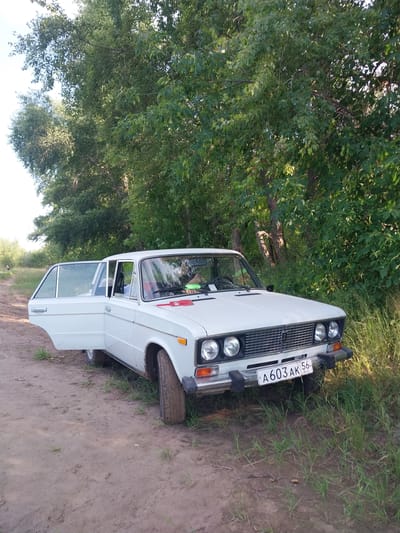 The image shows a white sedan parked on a dirt road next to a dense green foliage of trees and grass. The car's front passenger door and rear passenger door are open. A license plate reading "A603AK 56 RUS" is visible on the front of the car. The sun is shining, casting shadows on the scene. The environment appears to be rural or a park area on the outskirts of the city of Chkalov, Russia. There are no visible people in the immediate vicinity of the car.