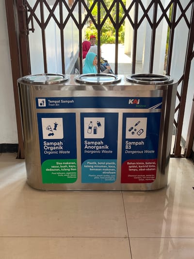 A three-compartment, stainless steel trash receptacle is centrally located in the foreground of the image. Each compartment is clearly labeled with text and icons in Indonesian and English, indicating different types of waste: "Sampah Organik" (Organic Waste), "Sampah Anorganik" (Inorganic Waste), and "Sampah B3" (Dangerous Waste). The receptacle is positioned on a tiled floor, and a metal, collapsible gate is visible behind it. Through the gate, a shaded outdoor area with white pillars and lush greenery can be seen. In the background, several individuals are visible. One person wearing a red headscarf and a red cap is smiling, while another person in a turquoise headscarf is looking to the side. The overall setting suggests an outdoor public space, possibly a transportation hub or a communal area in a place like Desa Tambakreja, Indonesia, given the visible signage and cultural context.