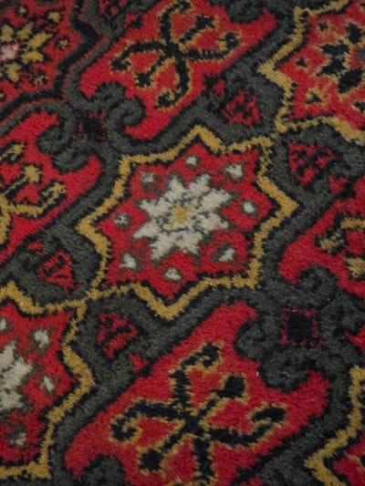 The image is a close-up, high-angle shot of a carpet with a complex geometric pattern. The dominant colors are red, black, and yellow, with some areas of white or cream. The pattern features intricate, repeating motifs that appear to be floral or star-shaped, outlined in black and yellow, and set against a red background. The texture of the carpet is visible, showing the woven fibers. The image is devoid of any context related to Petrushenko, Russia, or any discernible people, actions, or interactions. It is purely a visual study of the carpet's design and texture.