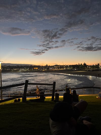 This image depicts an evening scene overlooking Bondi Beach from a grassy elevated vantage point in North Bondi, Australia. The sky transitions from a deep blue overhead to an orange and yellow glow at the horizon, with scattered clouds.
In the immediate foreground, a person is partially visible, reclining on green grass, wearing a dark shirt and light shorts, with their legs extended. Slightly further back, a couple is seated on the grass, facing the beach. To their left, a wooden post and rail fence runs horizontally, separating the grassy area from a lower paved promenade. A bright street light on a pole is positioned on the far left, emitting a strong yellow-white light.
On the promenade beyond the fence, a person wearing a plaid shirt walks from left to right. Below the promenade, the dark waters of Bondi Beach extend across the midground, with gentle waves breaking on the shore. Several indistinct figures are visible on the wet sand and in the shallow water along the beach.
In the background, the curving coastline of Bondi Beach leads to the illuminated urban landscape of Bondi. Numerous buildings are visible, illuminated by artificial lights. Streetlights line the distant promenade and roads. Several dark, tall trees, consistent with Norfolk Pines, are visible along the distant coastline. Construction cranes are silhouetted against the twilight sky on the distant horizon.
