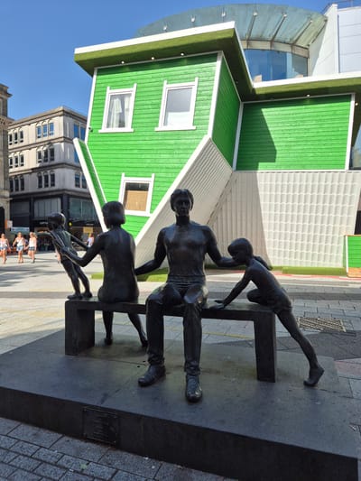 The image depicts a street scene in Cardiff, United Kingdom. In the foreground, there is a bronze statue of three people interacting with a set of uneven parallel bars. Behind the statue, there is a building with a prominent, bright green exterior and white trim, resembling an upside-down house. Pedestrians can be seen walking along the sidewalk behind the statue. Other buildings and businesses are visible in the background under a clear, blue sky.