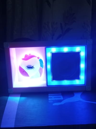 The image shows a close-up of a decorative item with two compartments, illuminated by soft, colored lights. The left compartment contains a small, white unicorn figurine with pink accents and closed eyes, lit by a soft pink glow. The right compartment is a dark square, outlined by bright blue LEDs. The item is placed on a surface that appears to be a dark blue box with a white Puma logo. The background consists of dark curtains with a floral pattern, dimly visible behind the object. The overall scene is dark with the primary light sources being the illuminations of the decorative item. There is no indication of the specific location in the city of Yampil, Ukraine, as the image is a close-up of an indoor object.