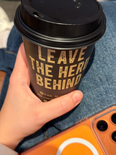 This close-up image depicts a person holding a black disposable coffee cup, identified by the prominent gold distressed text "LEAVE THE HERD BEHIND" and smaller branding "@ BLACK SHEEP" on its side. The cup has a black plastic lid with a visible drinking aperture. A light-skinned hand, with the thumb and index finger visible, grips the cup. The hand and cup rest against a surface of blue denim fabric, indicating the person is wearing jeans. To the lower right, an orange translucent smartphone case is partially visible, featuring a white circular Magsafe ring and multiple black camera lenses. The background is softly blurred, suggesting an indoor setting. The scene is situated in Bretton, United Kingdom.