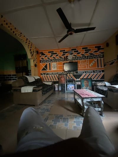 This image depicts the interior of a living room in Uffah, Nigeria, viewed from a first-person perspective with the observer's legs, clad in light-colored trousers, in the foreground. The room exhibits dim artificial lighting.
The walls are predominantly decorated with bold, geometric patterns in black and orange, accented with white. Specific patterns include brick-like designs, Greek key motifs, and abstract shapes. A white "GUCCI" logo is prominently painted on the orange section of the main wall, above which hangs a small, light-colored clock. Further to the right, an "adidas" logo is incorporated into the wall art. On the left side of the room, an arched doorway is visible, also adorned with geometric patterns. The ceiling is white, paneled, and features a black three-bladed ceiling fan. The floor is tiled with a repeating geometric pattern in shades of brown, grey, and possibly light beige.
Furnishings include two sofas or couches, positioned on either side of the room. The sofa on the left is dark-patterned with white cloths draped over its back and armrest. A central coffee table with a light, possibly distressed or marbled finish and dark legs occupies the middle ground. A pink or red fabric runner is placed on top of the coffee table, and a metallic cup rests on its lower shelf.
A television, appearing to be an older model (CRT or early flat-screen), is mounted on the main wall. Below the television, a stand holds several framed photographs, including one of a man in a suit with a beard. Another large framed portrait of a man, possibly in a formal setting, is prominently displayed on the floor to the right of the TV stand. Additional smaller framed photographs and decorative items are visible on surfaces throughout the room.