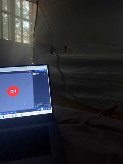 The image shows the interior of a space resembling a tent or canopy, possibly a makeshift structure within a room. A laptop is in the foreground, displaying a video conferencing interface with a red circular icon and two Cyrillic letters. A white window frame with an outside view of trees is visible behind the tent. There are fairy lights along the top of the window. A white bed with a figure obscured in the shadows. The location of Punta Gorda Town, Belize is not confirmed by the image.