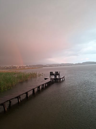 The image depicts a wooden pier extending into a dark, rain-swept lake in the Слабодкаўскі сельскі Савет, Belarus. The sky is overcast with a hint of pink and a faint rainbow is visible in the distance on the left side, suggesting a recent or ongoing rain shower. Reeds line the edge of the lake on the left, bordering the pier. At the end of the pier, two empty wooden chairs and a small table are positioned. The lake surface is disturbed by numerous raindrops, creating ripples. The far shore is lined with a dense forest and a cluster of buildings in the distance. The overall atmosphere is serene and melancholic, with the rain emphasizing the isolation of the scene.