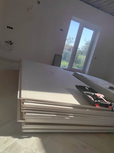 A stack of drywall sheets is leaning against a wall in an unfinished room. The sheets are white and appear to be new. In the background, a window looks out onto a sunny day with green trees and houses visible outside. The room itself is sparsely furnished, with electrical outlets visible on the wall. A black toolbox with red accents is resting on top of the drywall. The floor is made of light-colored wood planks. The overall impression is that of a construction or renovation project in progress. The lighting suggests it is daytime.