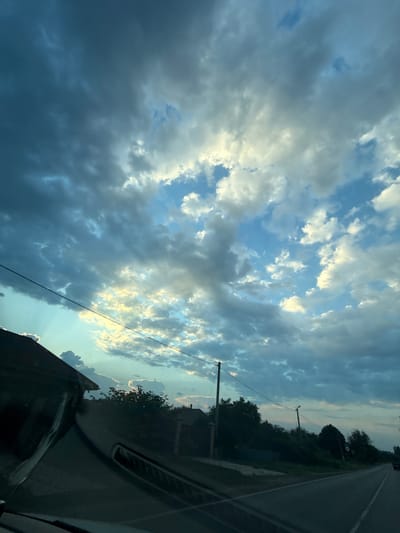 The image shows a view from inside a vehicle looking out at a rural landscape under a dramatic, cloudy sky. The sky is filled with a mix of dark, heavy storm clouds and brighter, illuminated clouds, suggesting either an approaching storm or a sunset/sunrise breaking through. Rays of light are visible filtering through the clouds on the left side of the image.
In the foreground, the dashboard of the vehicle is visible, with a dark, reflective surface. On the left, the corner of a building with a dark roof is visible, partially obscured by the vehicle's interior.
Beyond the vehicle, the landscape consists of a road stretching into the distance, with a tree line bordering its right side. Several power poles and lines are present along the road. A few buildings and structures are visible nestled among the trees. The overall environment appears to be a village or rural area, consistent with the location of Nizhneatamanskoe, Russia. No people are visible in the image.