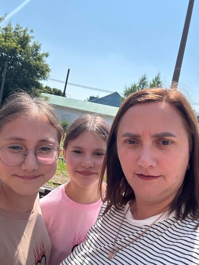 In this outdoor selfie, three individuals, presumably a mother and her two daughters, are captured from a low angle under a clear blue sky. The woman at the top appears to be in her late 30s or 40s, with shoulder-length brown hair and fair skin. She is wearing a black and white striped t-shirt and a delicate necklace with a circular pendant. Her expression is pleasant, with a slight smile.
Below her are two girls. The older girl, in the middle, has brown hair and a warm smile, wearing a pink t-shirt. The younger girl, in the foreground, is wearing glasses with round frames and a light brown t-shirt featuring a flame graphic. Both girls have cheerful expressions, suggesting a happy moment. The setting appears to be a casual outdoor environment, possibly a backyard or a park, with glimpses of green trees and a light-colored building in the background. The bright sunlight suggests it is daytime, likely mid-morning or afternoon. The overall mood of the image is joyful and familial. There is no visible text in the image.