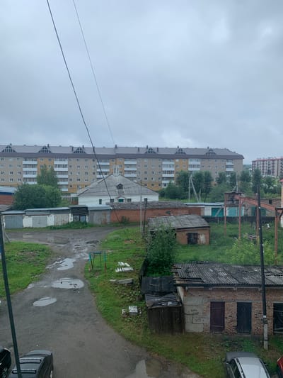 The image depicts a view of an overcast urban residential area in Volchansk, Russia. In the foreground, a dirty, partially paved path is visible, with several puddles of water scattered across it. To the left of the path, two cars are parked. Further into the scene, a row of garages with corrugated metal roofs and brick bases can be seen, some of which appear dilapidated. Behind these garages, a white building with a domed roof is situated. Dominating the background is a long, multi-story apartment building with a grey, pitched roof and light-colored facade, featuring numerous windows and balconies. Adjacent to this apartment building, another, taller apartment building with a red facade and white balconies is visible. The surrounding environment is characterized by green grass, trees, and electrical wires crisscrossing the sky, contributing to a somewhat neglected, post-industrial atmosphere. There are no people visible in the image.