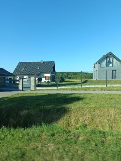 In Jabłonowo, Poland, two modern residential houses are visible under a clear blue sky. The house on the left has a dark gray tiled roof with dormer windows and is flanked by a gray fence and a gate constructed from concrete pillars and horizontal metal bars. The house on the right is a gray, two-story structure with a dark roof, featuring a large, triangular window with white curtains on its upper facade and a dark rectangular door on the lower level. A wire fence encloses the property. In the background, a dense green forest stretches across the horizon. The foreground consists of a mowed green lawn with some dry patches, and a paved walkway runs between the lawn and the fence. The image appears to have been taken from a moving vehicle, as evidenced by the motion blur in the grassy foreground.