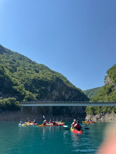 In the city of Bezuje, Montenegro, a group of kayakers navigate the clear, turquoise waters of a lake, framed by steep, tree-covered mountains under a bright blue sky. A large bridge spans the lake in the background, its structure prominent against the verdant slopes. The kayakers, dressed in various brightly colored life vests, are actively paddling, some in single kayaks and others in tandem. The scene depicts a leisurely outdoor activity in a picturesque, natural setting.