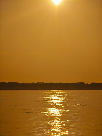 The image shows a wide body of water, possibly a river or lake, reflecting the warm, golden light of a setting sun. The sky is a gradient of orange and yellow, with the sun itself a bright, diffused orb near the top center of the frame, casting sunbeams downwards. In the distance, a dark, silhouetted treeline stretches across the horizon, with a few faint structures or lights visible within it. The water is rippled, creating a shimmering effect as the sunlight reflects off its surface. A lone bird appears to be flying low over the water near the treeline. The overall impression is one of a peaceful, natural landscape at dusk. The prompt requests information about Lesosibirsk, Russia, but this information is not present in the image itself and cannot be inferred from the visual content.