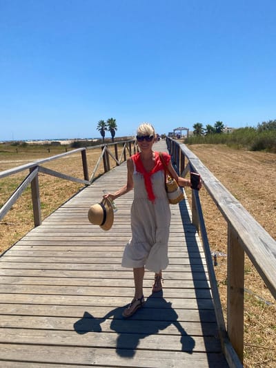 A woman walks on a wooden boardwalk toward the camera, holding a straw hat in her left hand and a smartphone in her right. She wears sunglasses, a white dress with a red scarf draped over her shoulders, and sandals. Her straw bag hangs from her left shoulder. The boardwalk, flanked by a wooden railing, is surrounded by dry grass and sparse vegetation, with palm trees visible in the background. The scene appears to be located in Conil de la Frontera, Spain, likely near a beach, as suggested by the presence of beach umbrellas and buildings in the distance. The sky is clear and blue, indicating a sunny day.