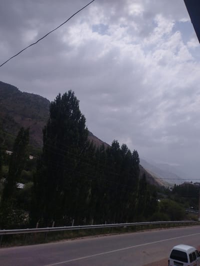 A grey, cloudy sky looms over a mountainous, rural landscape in Chaknak, Uzbekistan. A paved road runs horizontally across the bottom of the frame, with a metal guardrail separating it from a grassy verge. Tall, dark green trees line the hillside behind the road, obscuring much of the terrain. In the distance, the slopes of the mountains are visible, covered in sparse vegetation. A white van is parked on the side of the road in the lower right corner of the image. The overall scene is overcast and appears to be a quiet, natural setting.