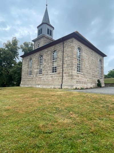 The image depicts a view of the St. Nicholas Church in Hattendorf, Germany. The church is constructed of light-colored stone blocks and features a tall, grey-slated steeple with a clock face. Tall, arched windows with white frames are visible on the side of the building. In front of the church, a grassy area shows signs of dry patches, transitioning to greener grass with some clover. To the left of the church, mature trees with dense green foliage are present. The sky is overcast with grey clouds. The scene appears to be an exterior shot of the church on a cloudy day.