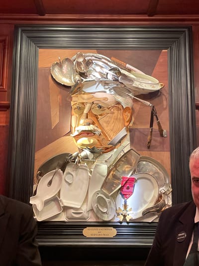 A framed, three-dimensional bust portrait of Auguste Escoffier, created by artist Bernard Pras using various kitchen utensils and dining ware, is prominently displayed on a dark reddish-brown wooden paneled wall. The artwork, primarily composed of metallic silverware, white porcelain dishes, and wooden elements, depicts Escoffier with a mustache and spectacles, wearing a chef's hat made of silver spoons and ladles. His torso is formed by an arrangement of white plates and platters, including a fish-shaped dish and a large round silver tray, adorned with a red-ribboned medal resembling the Légion d'honneur. A carving fork and knife with wooden handles hang to the right of the portrait's head. Beneath the artwork, a small plaque identifies the subject and artist. The dark, multi-ridged frame of the artwork is bordered by dark wood paneling and decorative molding. Portions of two individuals are visible in the foreground: the shoulder of a person in a dark suit on the left, and the right side of a man's face and shoulder, wearing a dark suit, tie, and a "Martin Maître d'hôtel" name tag, on the right. This scene is located at Les Grands Buffets restaurant in Narbonne, France.