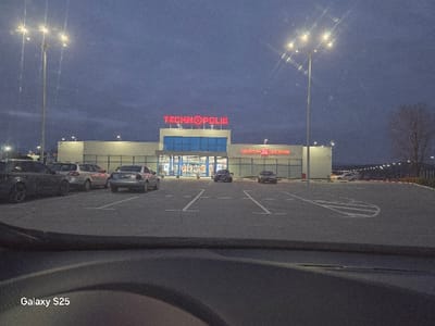 The scene depicts an evening view of a retail complex and its parking lot, identified as being in Novo Delchevo, Bulgaria. The primary commercial building, publicly associated with the Technopolis location in Gotse Delchev, Industrialna Zona, Bulgaria, features a light-colored facade with blue accents around its central entrance. A large, red, illuminated "TECHNOPOLIS" sign is mounted on the roofline above the entrance. To the right of the main entrance, another red, illuminated sign reads "МЕБЕЛИ 24 ВАСЕНОВ" (Mebeli 24 Vasenov). The parking lot is paved with asphalt and marked with white lines defining parking spaces and directional arrows.
Multiple vehicles are parked in the lot. In the foreground to the left, a dark-colored sedan or SUV is parked. Behind it, a light-colored SUV is visible, followed by a silver sedan. Additional vehicles are discernible further in the parking lot to the right. The area is illuminated by several tall streetlights, each featuring multiple LED fixtures, and by facade lighting on the retail building. The sky is dark blue, indicating dusk or night. No individuals are visible. The image is captured from inside a vehicle, with the dashboard visible in the lower foreground. A "Galaxy S25" watermark is present in the bottom left corner.