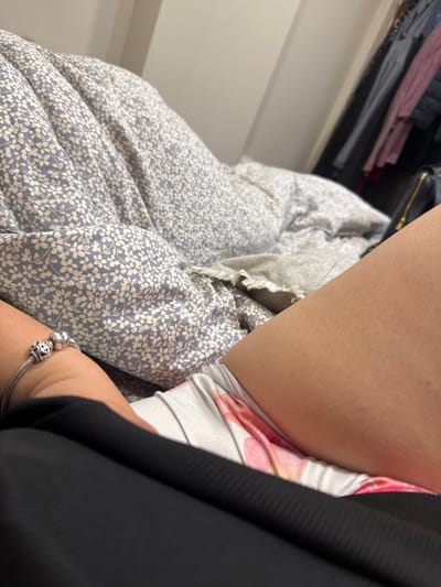 The image shows a close-up view of a person lying on a bed. The person is wearing a black garment and floral print underwear. A bracelet with charms is visible on the person's left wrist. The bed is covered with a blue and white floral patterned duvet and a gray blanket. In the background, a closet with hanging clothes is visible. The overall setting appears to be a bedroom in a home. There is no information in the image to suggest it is located in Petkovo, Bulgaria, or any specific city.