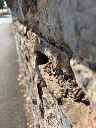 The image shows a close-up, low-angle, shallow depth of field photograph of a rough, textured stone wall. The wall is constructed of irregular, weathered stones in shades of grey, brown, and reddish-brown, with some patches of lichen. The stones are loosely fitted, with visible gaps and crevices, some of which appear to contain fine spiderwebs. The foreground focuses on a section of the wall where the texture is highly detailed, showing granular surfaces and sharp edges. To the left of the wall, a paved surface, likely a road or sidewalk, is visible and out of focus, with a subtle bokeh effect. The background is blurred, showing a hint of greenery and a structure, possibly a building, in the far distance. The lighting suggests it is daytime, with sunlight casting shadows and highlighting the textures of the stone. There are no people or animals visible in the frame. The location is identified as Torquay, United Kingdom.
