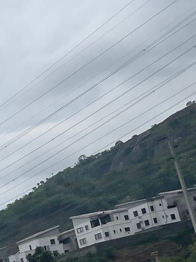 The image captures a tilted view of a landscape in Jiru, Nigeria, dominated by power lines crisscrossing the sky. Below the power lines, a steep, tree-covered hill forms the backdrop. In the foreground, a cluster of modern white buildings with dark rectangular windows is nestled on the hillside, suggesting a residential or commercial development. The sky is overcast and a light gray, contributing to a subdued atmosphere.