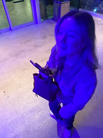 A woman stands on a concrete surface under blue lighting. She has long hair and is wearing a jacket or shirt. She carries a black bag in her left hand. Her face is visible, and she appears to be smiling. The background includes a window with a reflection of outdoor scenery. The image is taken from a high angle, giving a top-down perspective.