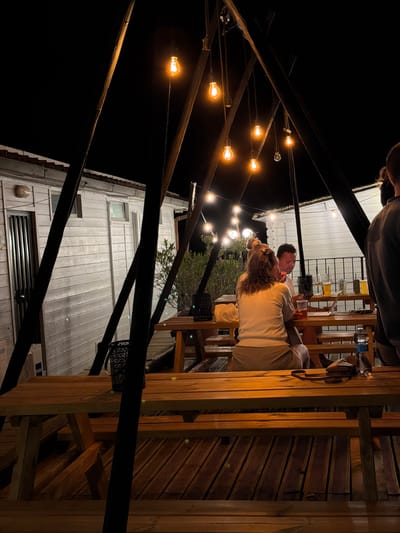 The image depicts an outdoor seating area at night, illuminated by string lights. In the foreground, a wooden picnic table and benches are visible. In the background, a white building with wooden siding and a door with a metal grate is present. Two individuals are seated at a table, looking at a laptop. Another person is partially visible on the right side of the image, with their back to the camera. There are several glasses of drinks on the table, as well as a bottle of water and a purse. The setting appears to be a casual outdoor dining or gathering space. The location is identified as Cavriglia, Italy.