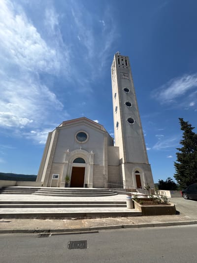 The image shows the Church of Christ the King in Neum, Bosnia and Herzegovina, viewed from a low angle. The church is a light-colored building with a tall, slender bell tower featuring circular windows. The facade has a large arched doorway and a circular window above. A wide set of steps leads up to the entrance. The sky is blue with scattered white clouds. A road and some greenery are visible on the right side of the frame. A tree is visible partially obscuring the church from the right.
