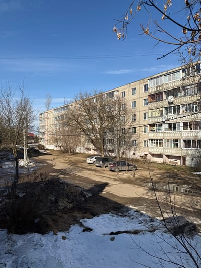 This image depicts an urban residential scene in Kurganovo, Russia, under a clear blue sky. A large, multi-story apartment building with a light-colored facade, featuring numerous windows and enclosed balconies, dominates the middle and right sections of the frame. One balcony on an upper floor displays a satellite dish. The ground level comprises an unpaved dirt and gravel area, serving as a parking lot, with visible patches of melting snow, mud, and water puddles. In the immediate foreground, a substantial mound of dirty, melting snow and earth is present. Three soiled passenger vehicles are parked along the apartment building: a white sedan, a dark-colored sedan, and another dark-colored sedan. Numerous deciduous trees, all bare of leaves, are scattered throughout the scene, with their branches partially obscuring the building and sky. Bare bushes are visible near the building's base. Overhead power lines traverse the upper portion of the image. The scene is illuminated by direct sunlight, casting distinct shadows. No individuals or discernible activities are visible.