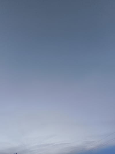 The image depicts a wide-angle view of a pale blue sky with a subtle gradient, transitioning to a lighter hue near the horizon. There are faint, wispy clouds scattered across the lower portion of the sky. In the far distance, along the bottom edge of the frame, faint outlines of distant structures and power lines are barely visible. The overall scene is one of an open, outdoor environment, likely during the daytime. No specific details about the city of Патоцкі сельскі Савет, Belarus, can be discerned from this image alone.