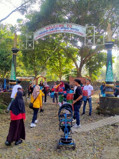 The image captures an outdoor scene at the entrance to Ngunut The Hot Spring Water, Sendang Panguripan, Sragen, Central Java, Indonesia, situated within the broader area of Jetis. A white, arched overhead sign with black lettering, stating "WELCOME TO NGUNUT THE HOT SPRING WATER SENDANG PANGURIPAN - SRAGEN JATENG," spans across a pathway. The sign is supported by two decorative pillars, each featuring a dark base and an upright, green, flame-like sculpture.
Approximately 15-20 individuals, predominantly adults, are visible on the unpaved path, which is covered with small rocks and scattered dry leaves. Many individuals are carrying or holding stacks of colorful plastic baskets or containers in red, green, blue, pink, and grey. Several women wear hijabs. In the immediate foreground, a blue child's tricycle or stroller is positioned, facing away from the viewer.
A man in a white polo shirt on the right holds multiple red baskets. To his left, a man in a black t-shirt also holds baskets. A group of individuals in the mid-ground, including several women, are similarly holding or handling these containers, suggesting an organized activity. A woman in a yellow long-sleeved shirt and a patterned hijab walks towards the left, away from the foreground. In the left foreground, a woman in a red shirt and black hijab walks directly away from the camera. The background consists of dense green foliage and large trees under clear daylight.