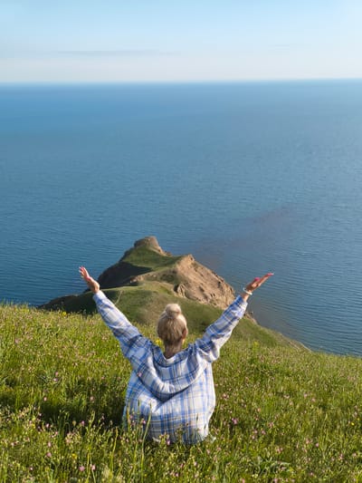 A full, high-angle shot depicts a woman seated on a grassy hillside, facing away from the viewer, with her arms raised in a posture of apparent joy or exhilaration. The scene is set in Коктебельский поселковый совет, Ukraine.
The woman, who appears to be Caucasian, has blonde hair pulled back in a bun. She is wearing a light blue and white plaid shirt or jacket. The hillside is covered in green grass and dotted with small wildflowers, primarily pink and yellow.
In the background, a striking body of water stretches out to the horizon, with the sky above a clear, pale blue. A landform of brown rock and green vegetation juts into the water, adding depth and interest to the landscape. The woman's pose and the expansive view suggest a moment of serenity and connection with nature.