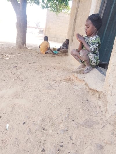 The scene depicts an outdoor area in the city of Kanwa, Nigeria, under bright daylight. In the foreground, a young child, approximately 4-6 years old, with dark skin and a black headwrap, squats barefoot on a beige concrete ledge against a building. The child wears a light-colored dress with a green floral or abstract pattern, and holds hands clasped together in front of the chest, with lips slightly parted. Dust and dirt are visible on the child's bare feet and lower legs. To the left in the midground, a second child, wearing a yellow shirt and light pants, sits on the dry, light-brown dirt ground beneath a tree, interacting with a light blue bowl and other small items on the ground. A third child, wearing a dark top, is seated further back against a light-colored wall, appearing to observe or manipulate items within a woven basket. The ground consists of dry, sandy earth mixed with scattered small rocks and dark debris. The background features light-colored mud-brick or concrete block structures, with a dark green door partially visible behind the foreground child.